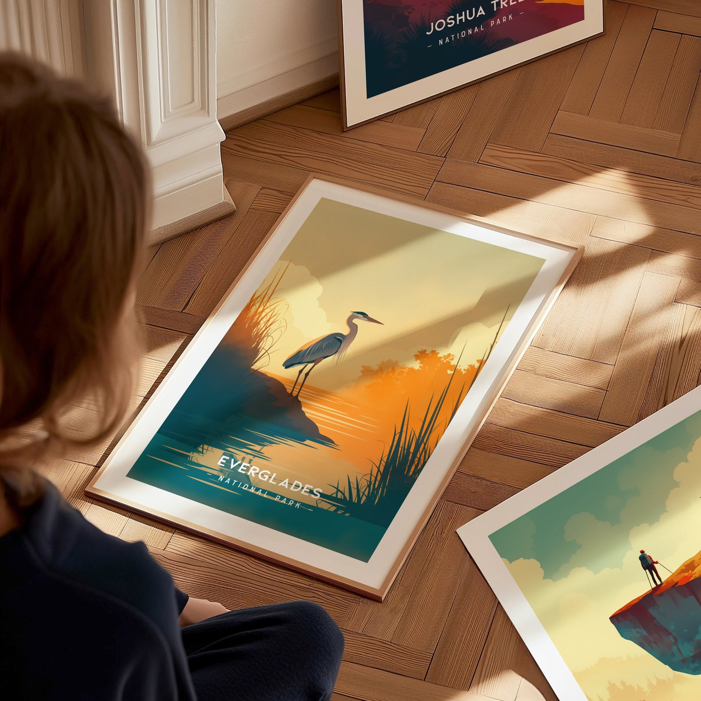 A person sits on a wooden floor, admiring a framed poster of the Everglades with a heron by a sunset-lit waters edge. Additional posters are partially visible.