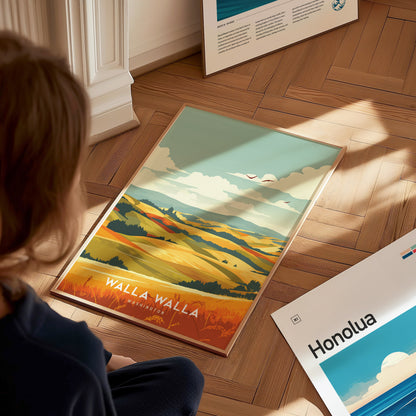 A person sits on a wooden floor, admiring a colorful Walla Walla Washington poster depicting rolling hills and vineyards under a blue sky. Nearby, another poster partially reads Honolua.