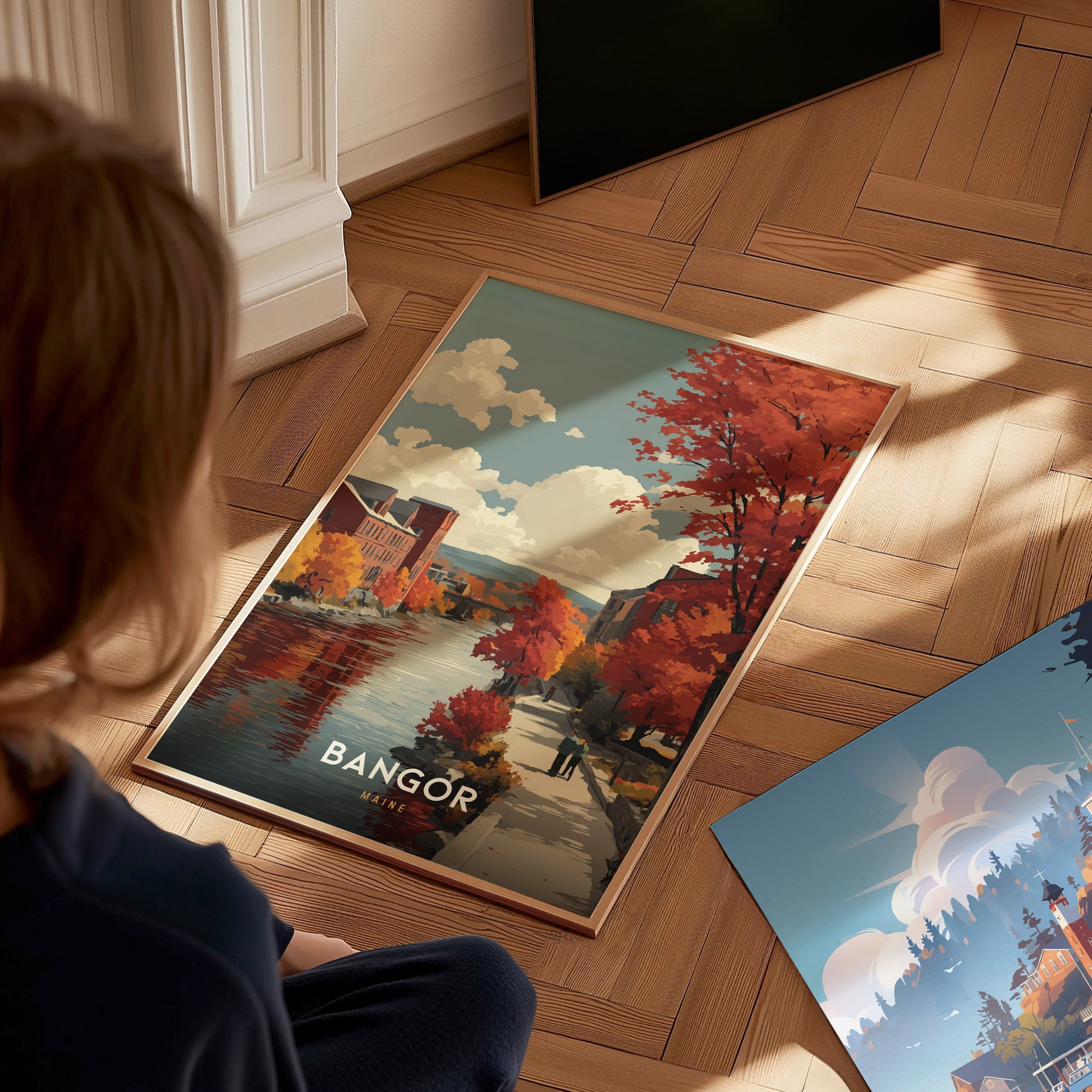 A person sitting on a wooden floor examines a colorful poster featuring a scenic view of a river surrounded by autumn trees with vibrant red and orange leaves.