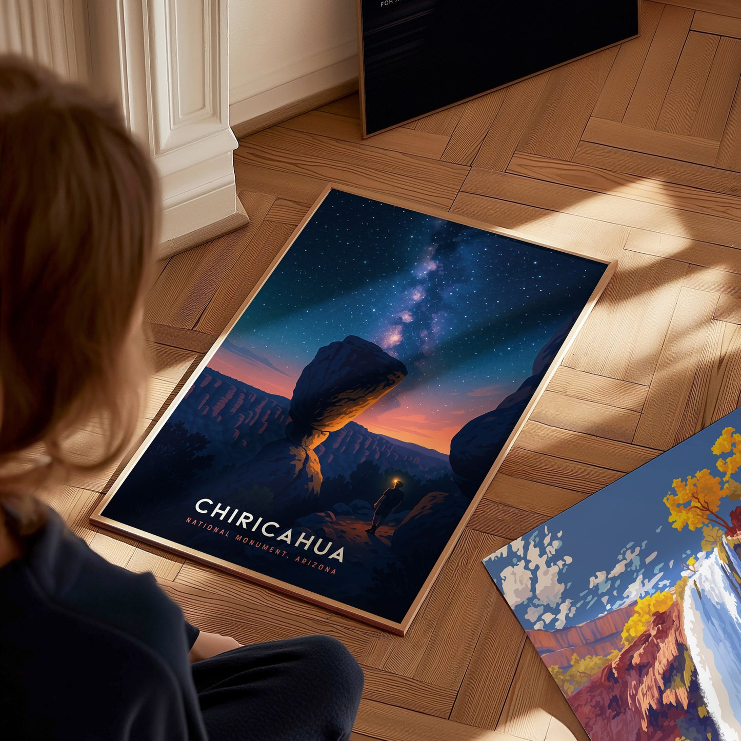 A child sits on a wooden floor, gazing at a vibrant poster depicting Chiricahua National Monument in Arizona. The artwork features a starry night sky above a dramatic rock formation, evoking a sense of wonder and connection to nature.