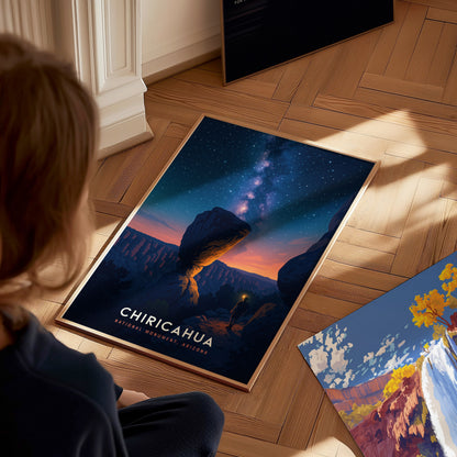 A child sits on a wooden floor, gazing at a vibrant poster depicting Chiricahua National Monument in Arizona. The artwork features a starry night sky above a dramatic rock formation, evoking a sense of wonder and connection to nature.