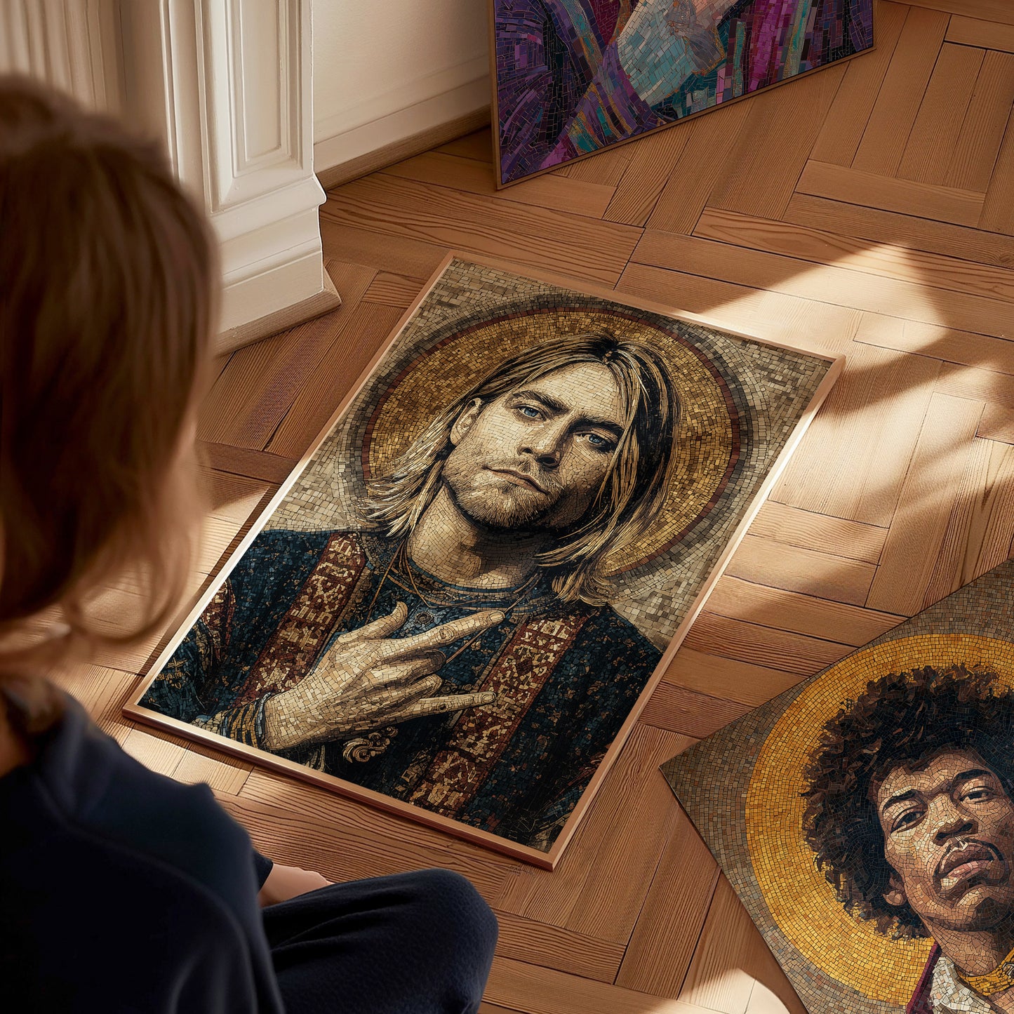 A person sits on a wooden floor, gazing at a mosaic-style portrait of Kurt Cobain, featuring intricate details and a halo effect. Nearby, additional framed artworks of other musicians can be seen, adding to the artistic atmosphere.