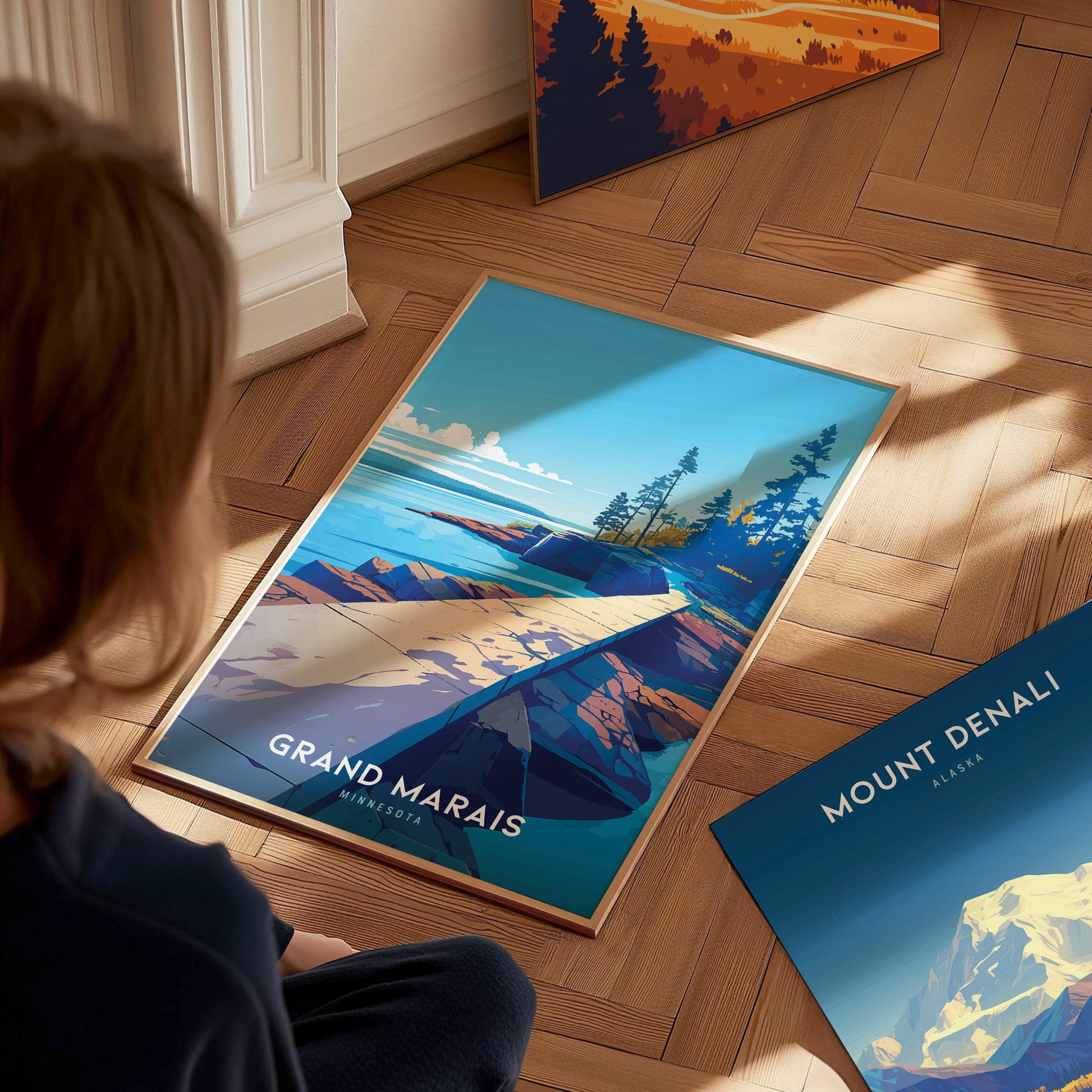 A person sits on a wooden floor observing a colorful framed poster of Grand Marais, Minnesota, depicting a coastal landscape with trees and rocky shores.