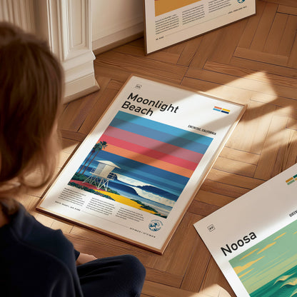 A person sits on a wooden floor, looking at several vibrant beach posters. One poster features Moonlight Beach with bold stripes in warm colors, a lifeguard tower, and a coastal scene depicting waves and palm trees.