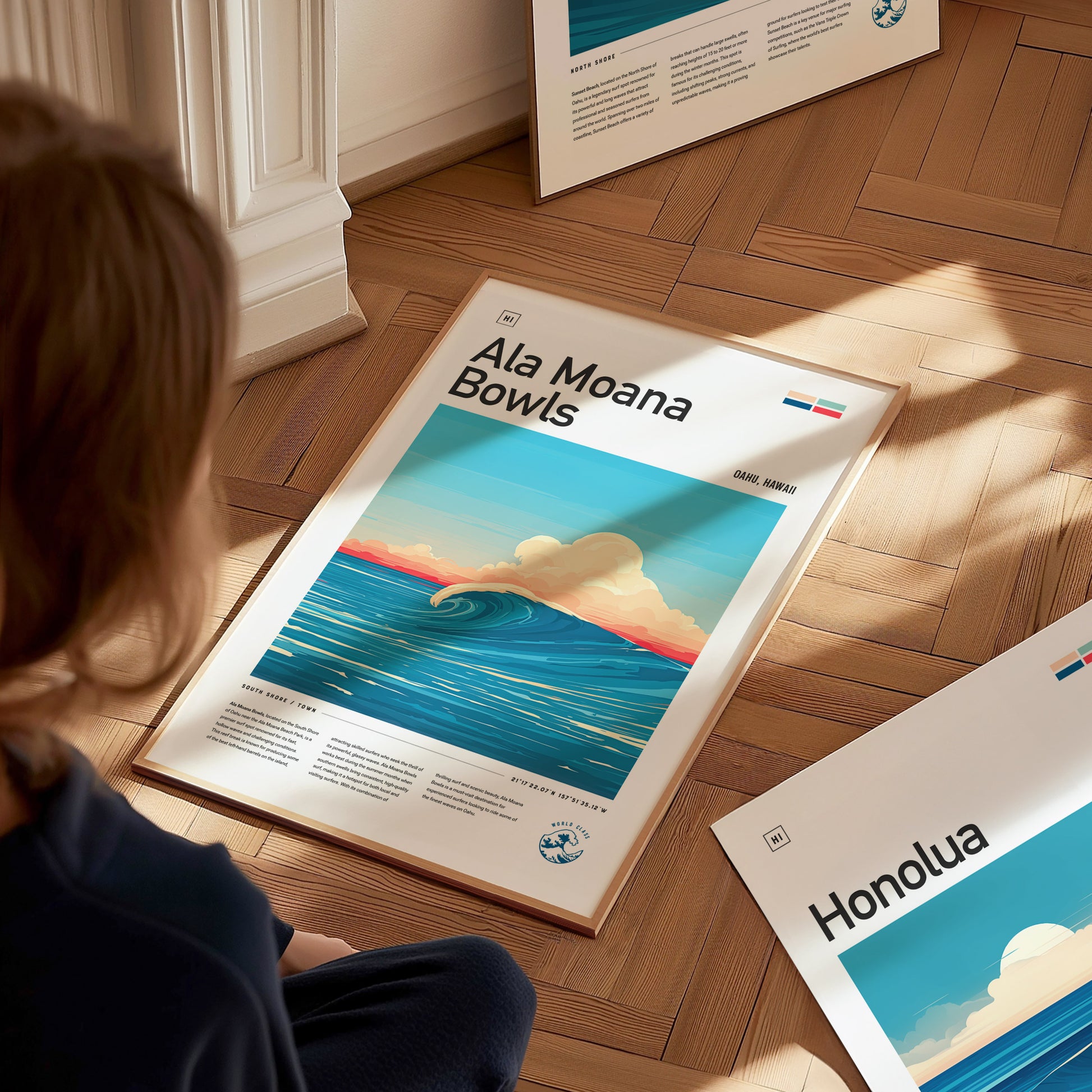 A person sits on a wooden floor, viewing a framed Ala Moana Bowls poster featuring a stylized ocean wave and colorful sky. Another poster labeled Honolua is nearby.