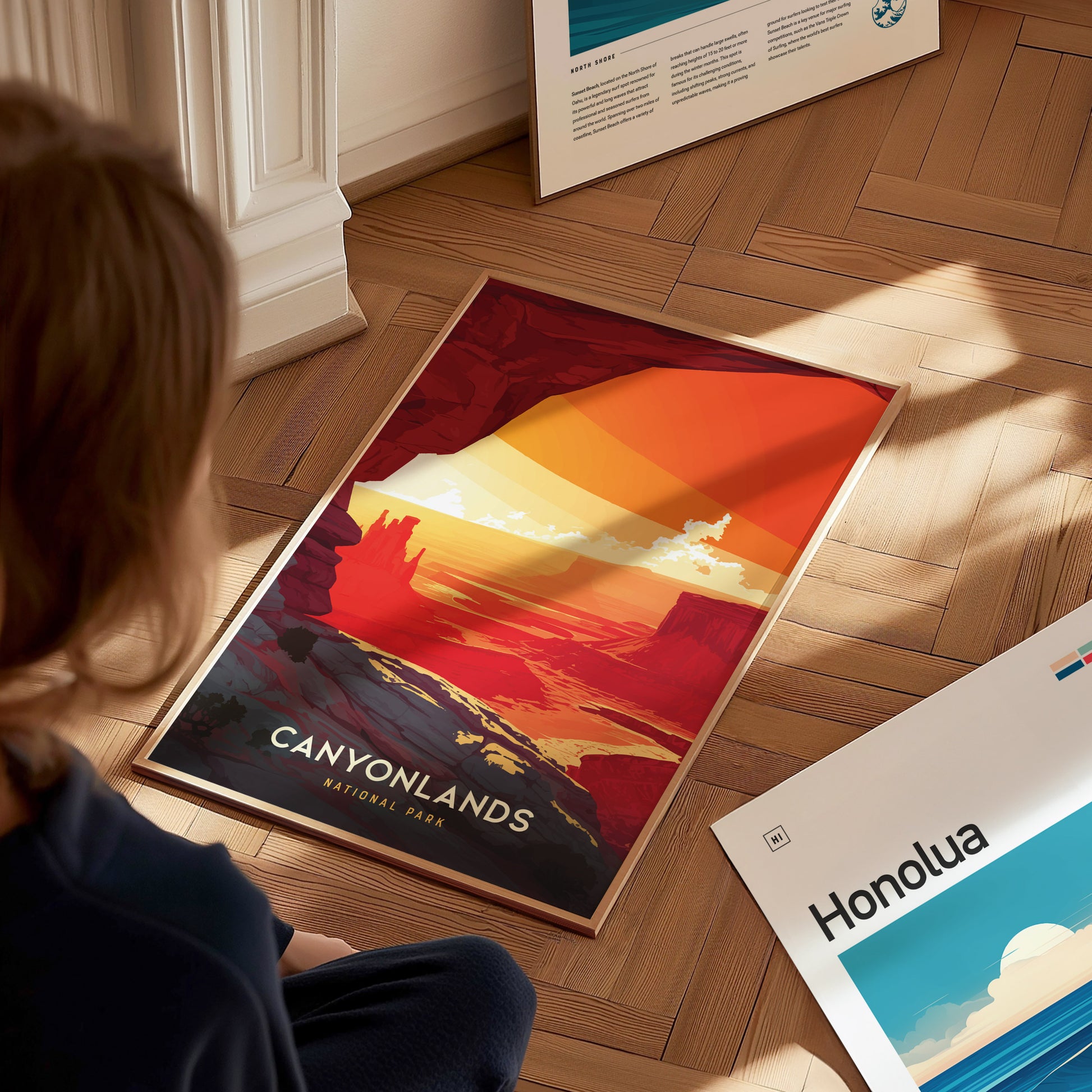 A person is viewing a vibrant Canyonlands National Park poster on a wooden floor, featuring bold reds and oranges depicting desert canyons. Nearby, another artwork titled Honolua lies flat.
