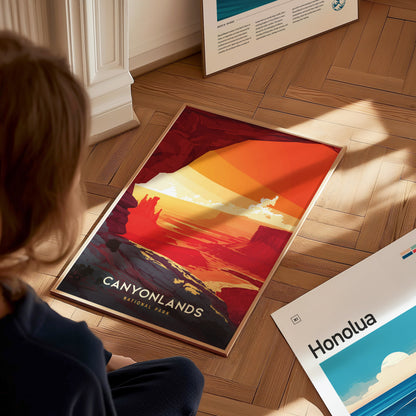 A person is viewing a vibrant Canyonlands National Park poster on a wooden floor, featuring bold reds and oranges depicting desert canyons. Nearby, another artwork titled Honolua lies flat.