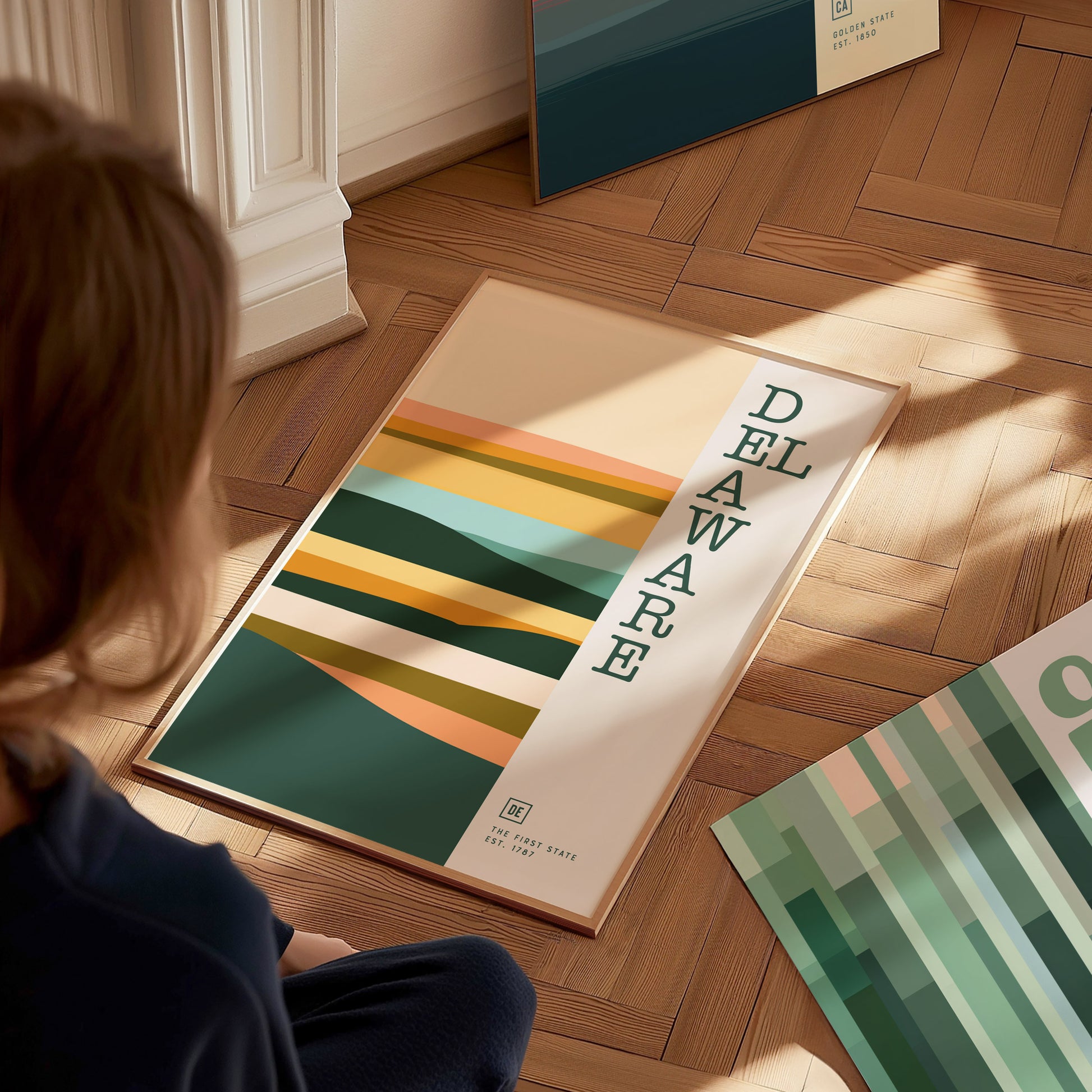 A person sits on a wooden floor, looking at a framed Delaware poster featuring abstract, layered shapes in warm, earthy tones. Another similar poster is partially visible nearby.
