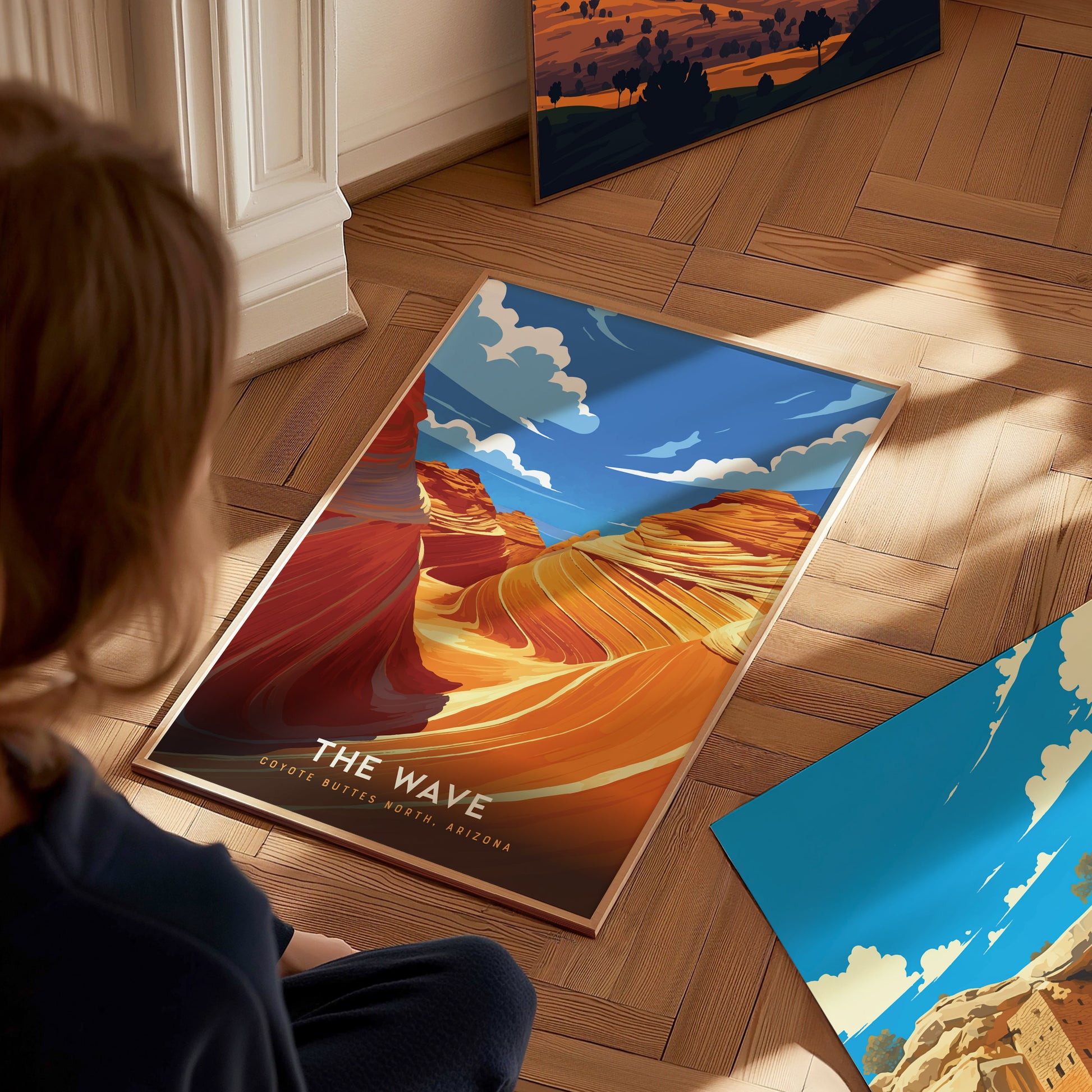 A person views a colorful poster of The Wave in Coyote Buttes, Arizona, featuring swirling sandstone formations under a vivid blue sky, placed on a wooden floor.