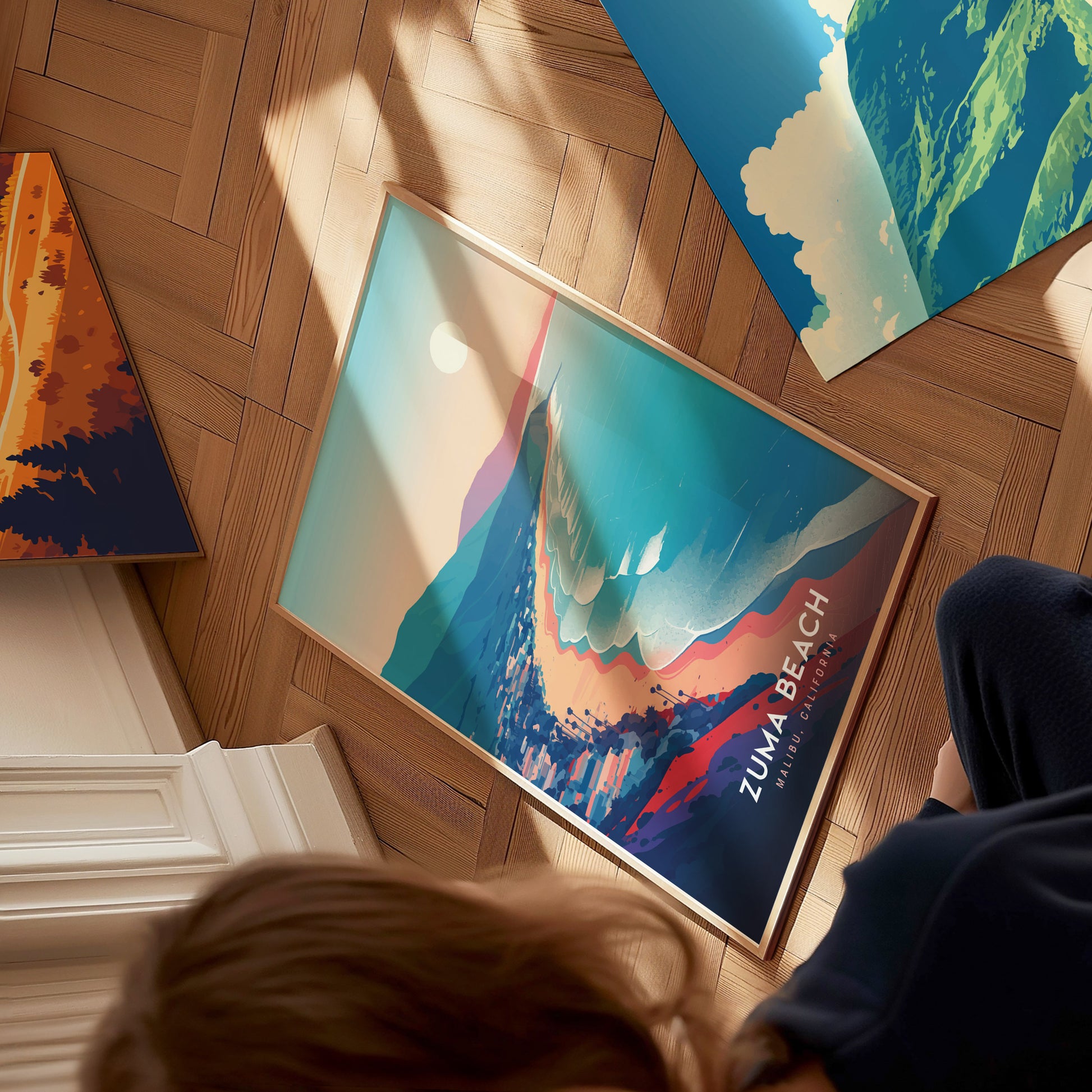 A person is sitting on a wooden floor, looking down at a colorful art print titled “Zuma Beach.” The print features vibrant waves and a stylized sun, with mountains in the background, creating a serene beach scene.