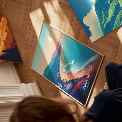 A person is sitting on a wooden floor, looking down at a colorful art print titled “Zuma Beach.” The print features vibrant waves and a stylized sun, with mountains in the background, creating a serene beach scene.