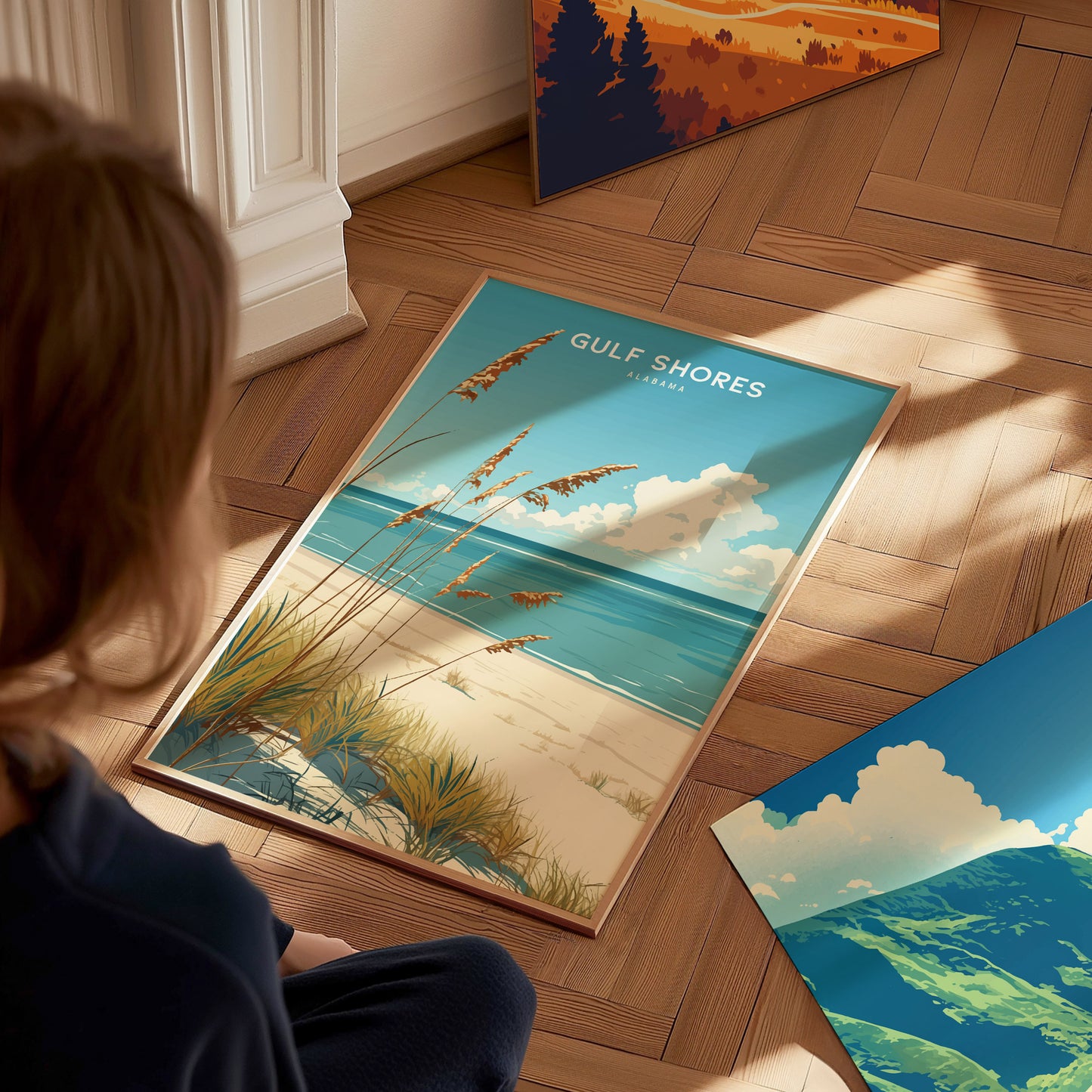 A person sits on a wooden floor, looking at a framed Gulf Shores, Alabama beach poster featuring sandy shores and a serene ocean under a bright blue sky.