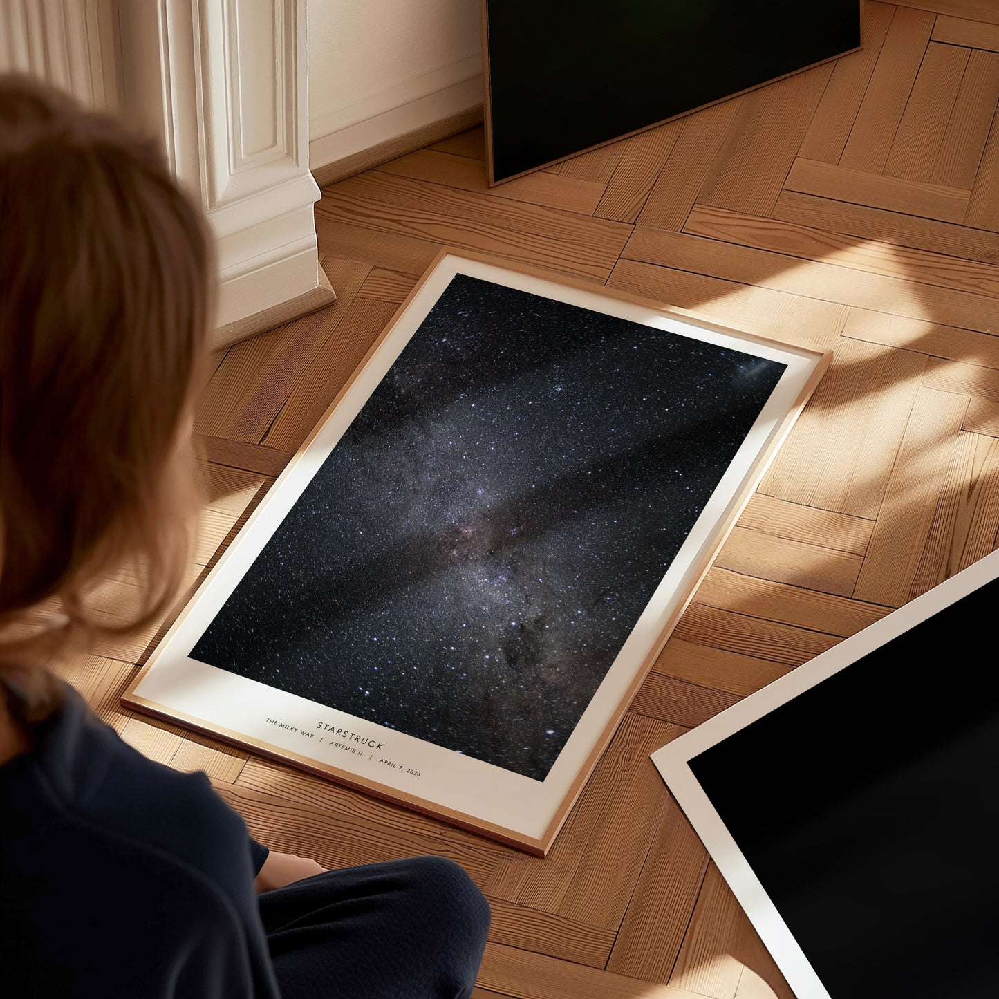 A child sits on a wooden floor, gazing at a framed poster featuring a stunning view of a starry night sky filled with countless stars and a hint of a galaxy. The warm light from the side enhances the tranquil atmosphere of the scene.