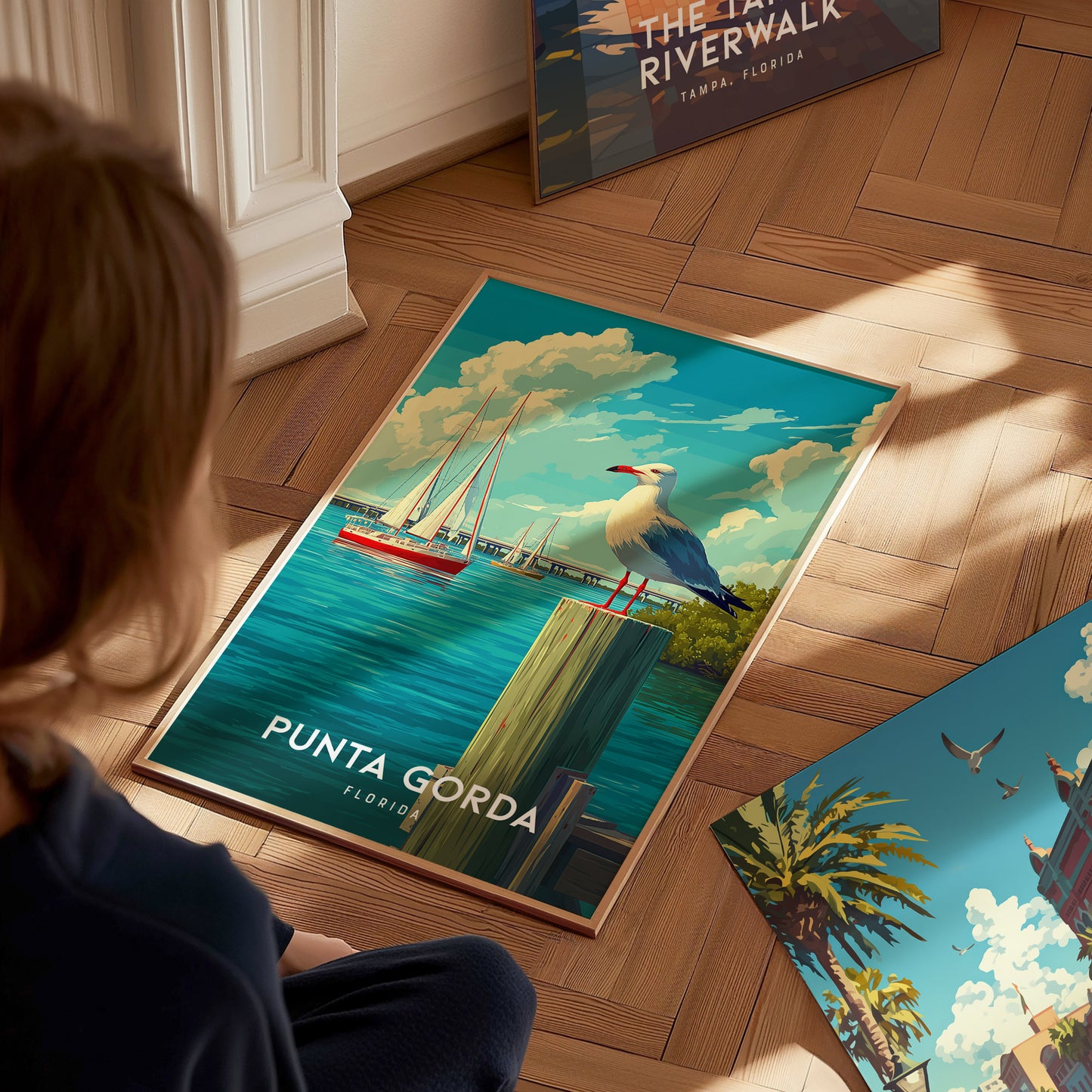 A child sits on a wooden floor, gazing at a vibrant poster featuring a coastal scene of Punta Gorda, Florida. The artwork depicts a white bird perched on a wooden post, with sailboats in the background and a clear blue sky above.