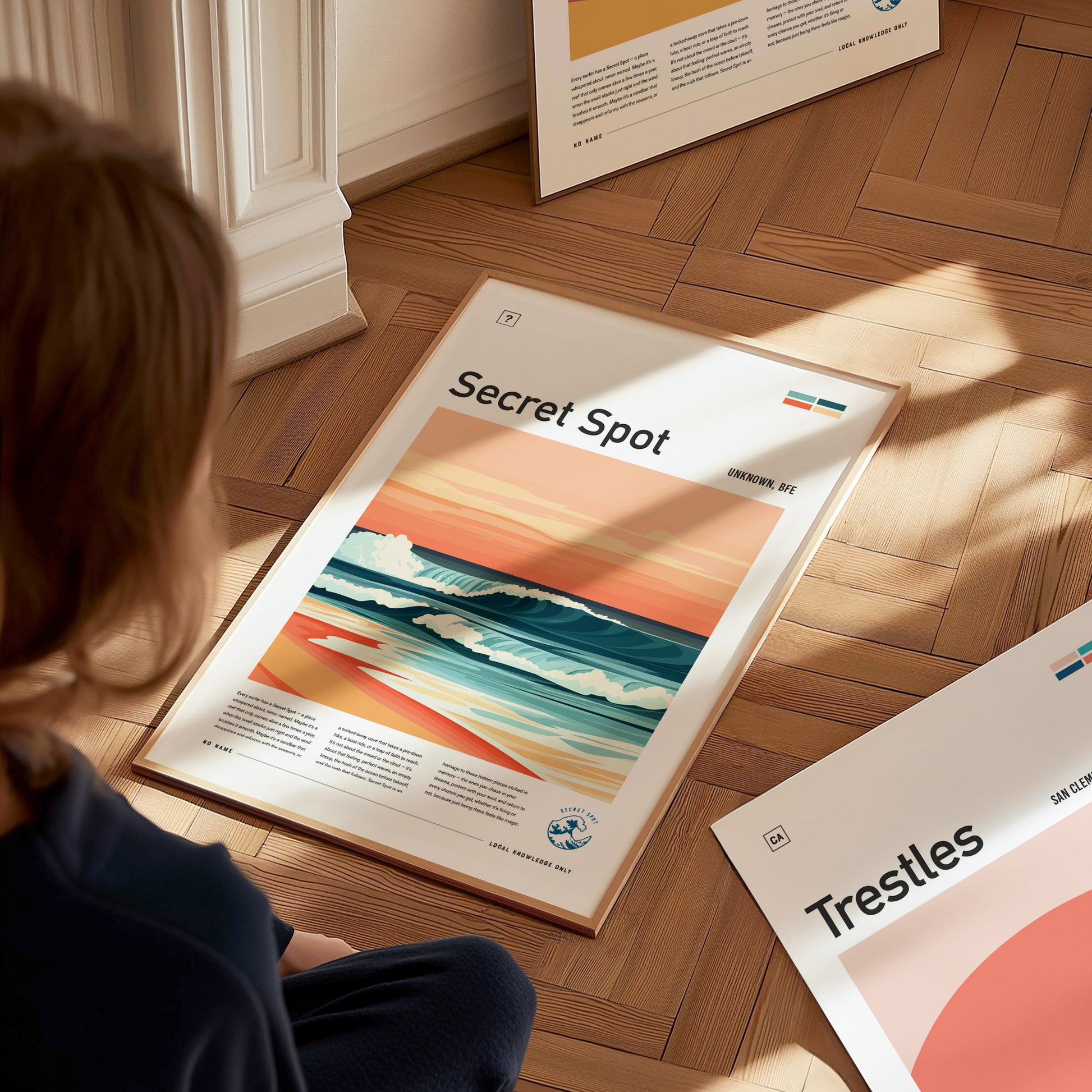 A person sits on a wooden floor, looking at a framed surf poster titled Secret Spot featuring a stylized wave under pastel skies. Other posters are partially visible nearby.