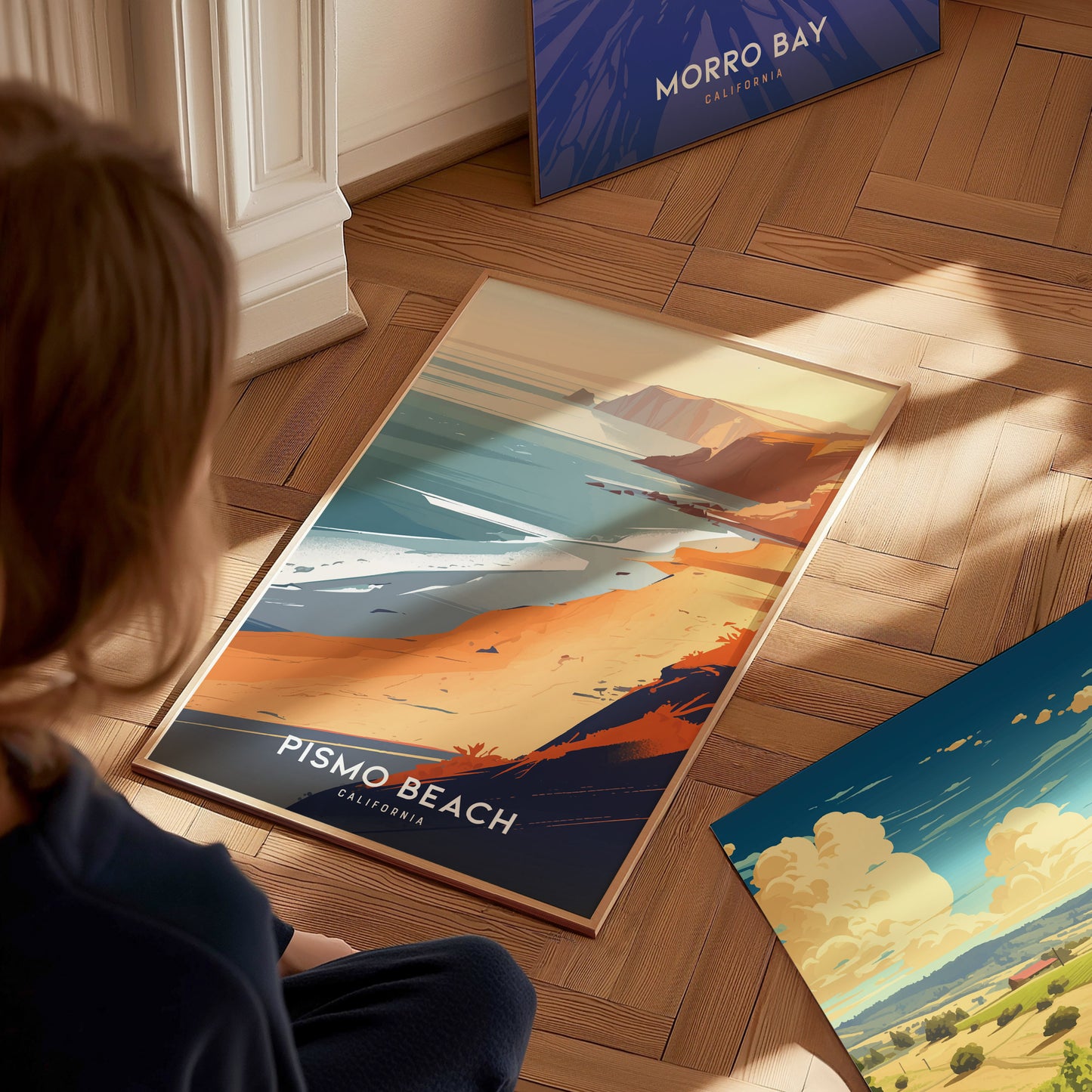 A person sits on a wooden floor admiring a Pismo Beach poster, depicting a scenic coastline with cliffs and ocean waves. Nearby, a Morro Bay poster leans against the wall.