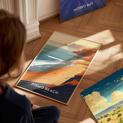 A person sits on a wooden floor admiring a Pismo Beach poster, depicting a scenic coastline with cliffs and ocean waves. Nearby, a Morro Bay poster leans against the wall.