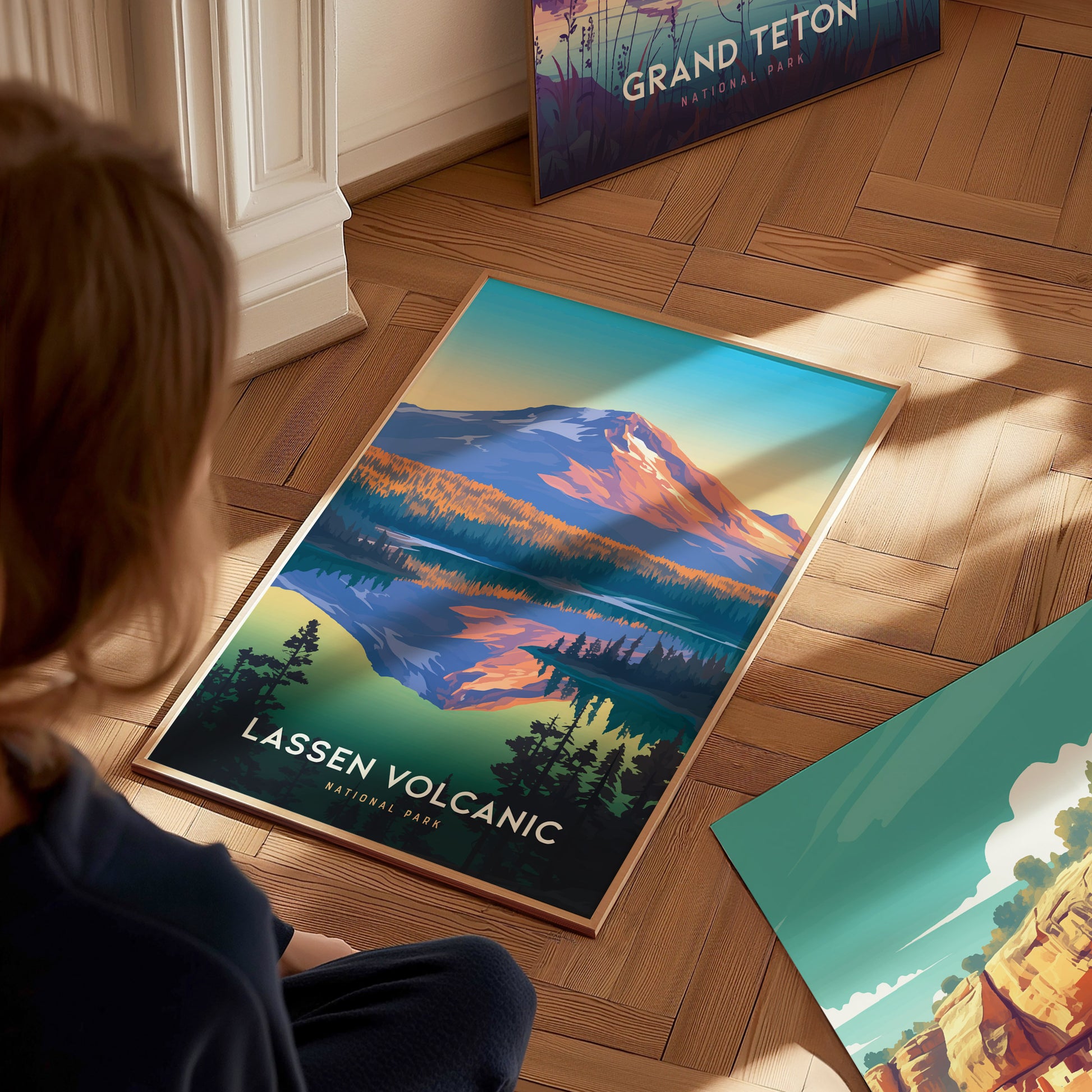 A person looks at a colorful Lassen Volcanic National Park poster on a wooden floor, showcasing a mountain landscape with trees and a lake. Another poster is partially visible nearby.