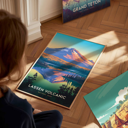 A person looks at a colorful Lassen Volcanic National Park poster on a wooden floor, showcasing a mountain landscape with trees and a lake. Another poster is partially visible nearby.