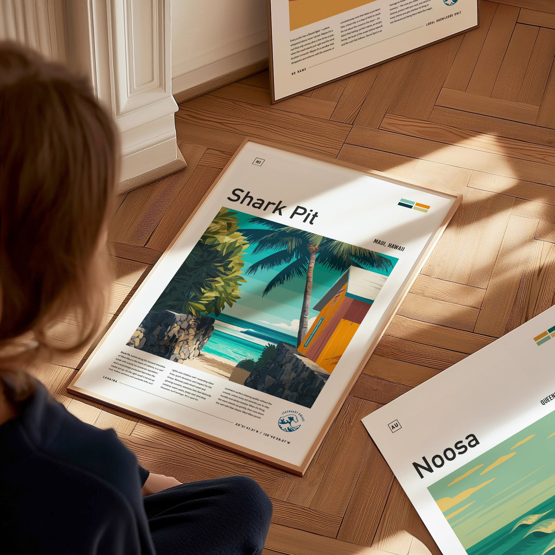 A child sits on a wooden floor, looking at framed art prints featuring beach scenes. One print titled Shark Pit showcases a tropical landscape with palm trees, a colorful beach hut, and turquoise waves under a clear sky.