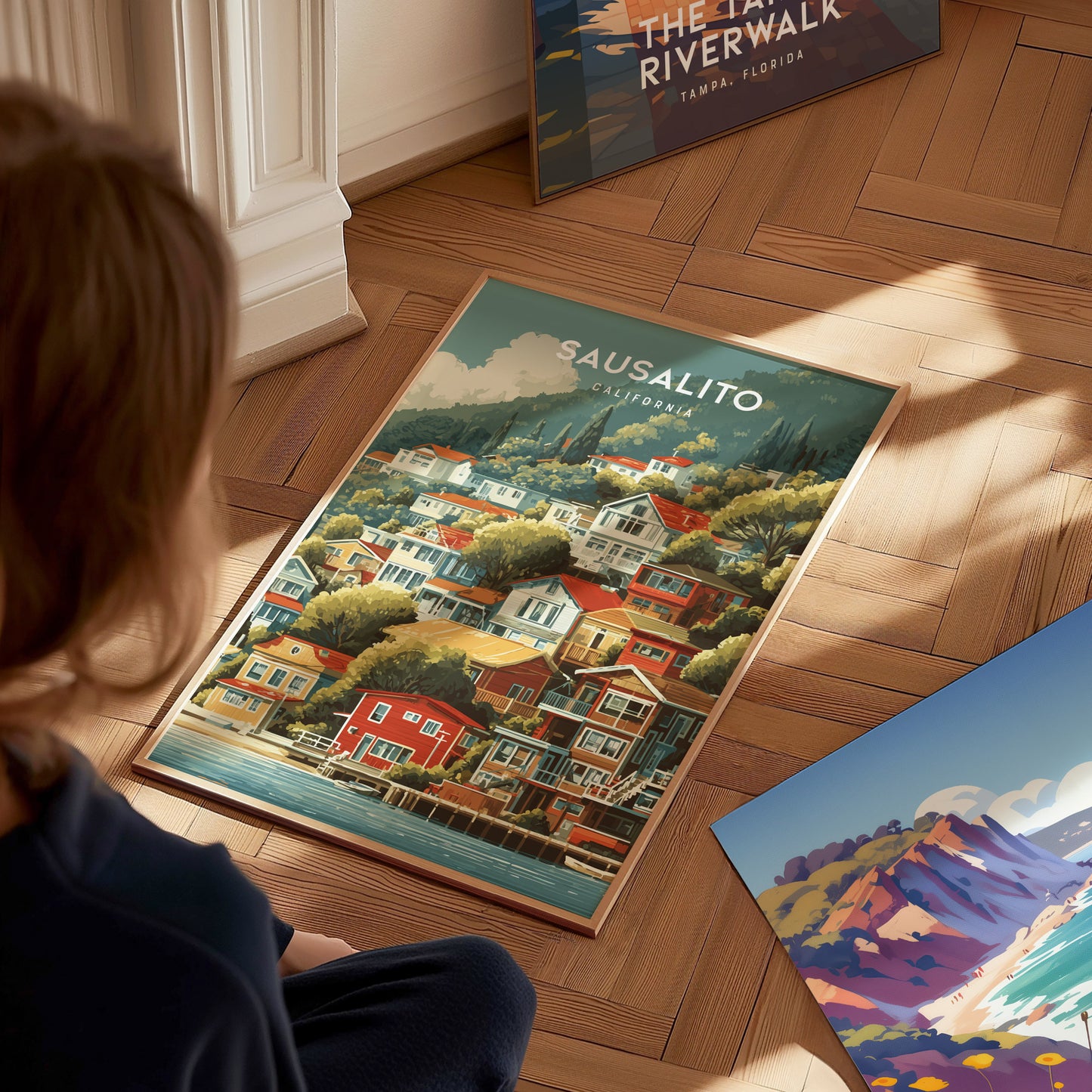 A child sits on a wooden floor, gazing at a colorful poster featuring a scenic view of Sausalito, California. The artwork showcases vibrant houses along the waterfront, surrounded by lush green hills and a clear blue sky.
