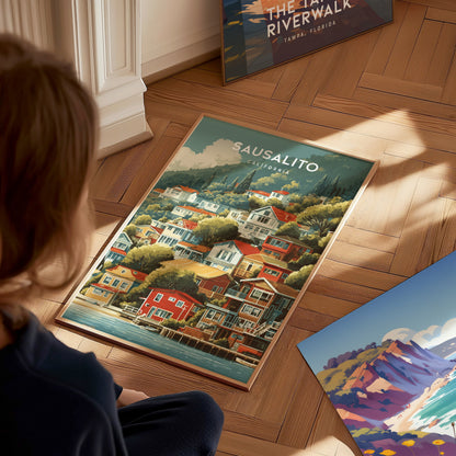 A child sits on a wooden floor, gazing at a colorful poster featuring a scenic view of Sausalito, California. The artwork showcases vibrant houses along the waterfront, surrounded by lush green hills and a clear blue sky.