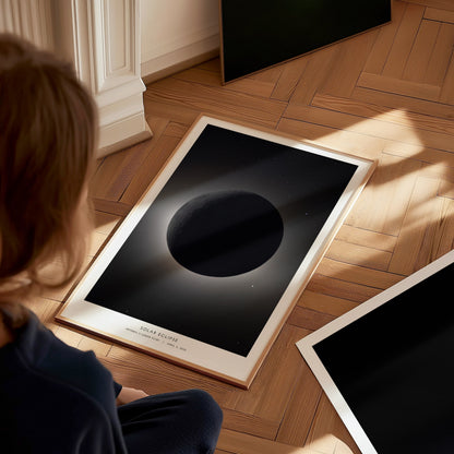 A child sits on a wooden floor, gazing at a framed poster depicting a dark celestial body with a soft glow, surrounded by a starry background. Nearby, an additional unframed poster lies flat on the floor, showcasing a solid black design.