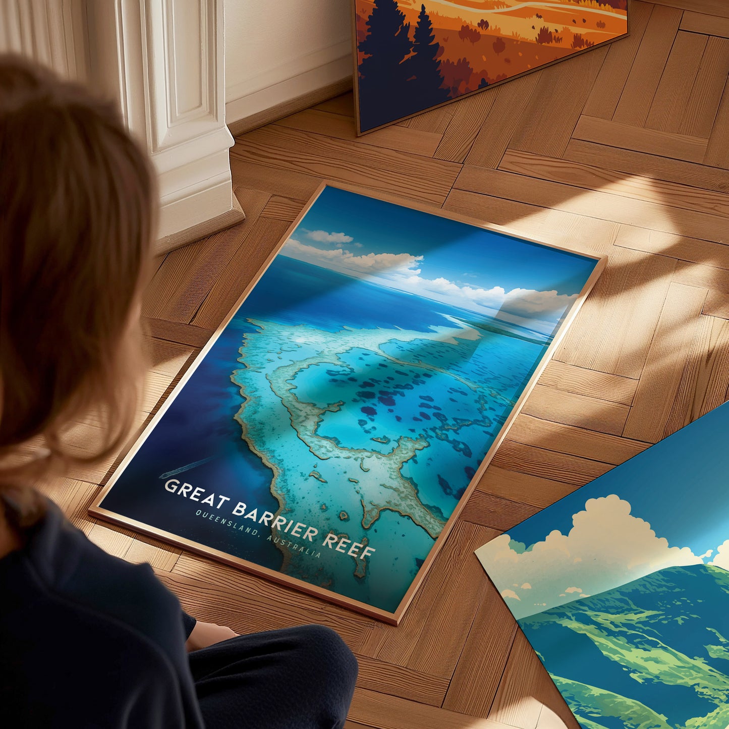 A person observes a vibrant Great Barrier Reef poster on a wooden floor, showcasing turquoise waters and coral formations. Other colorful posters are partially visible nearby.