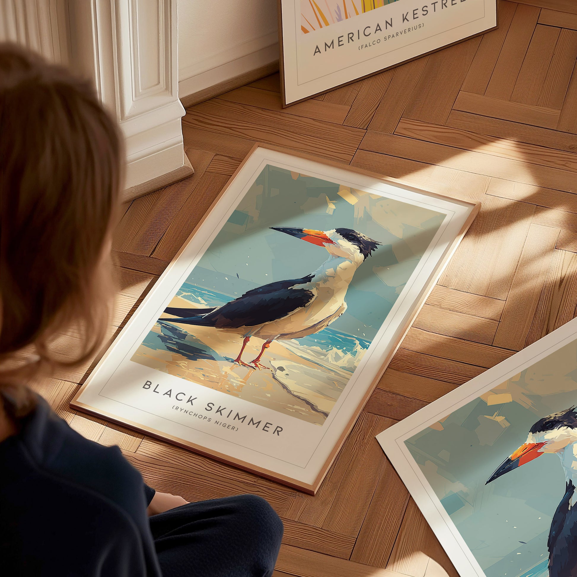 A young child sits on a wooden floor, gazing at a vibrant poster of a black skimmer, featuring the bird standing on sandy shores. The artwork showcases the skimmers distinctive bill and coastal habitat, framed beautifully.