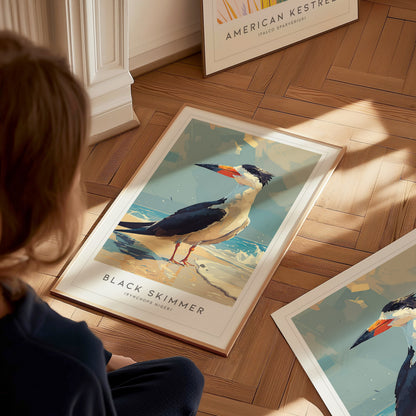 A young child sits on a wooden floor, gazing at a vibrant poster of a black skimmer, featuring the bird standing on sandy shores. The artwork showcases the skimmers distinctive bill and coastal habitat, framed beautifully.