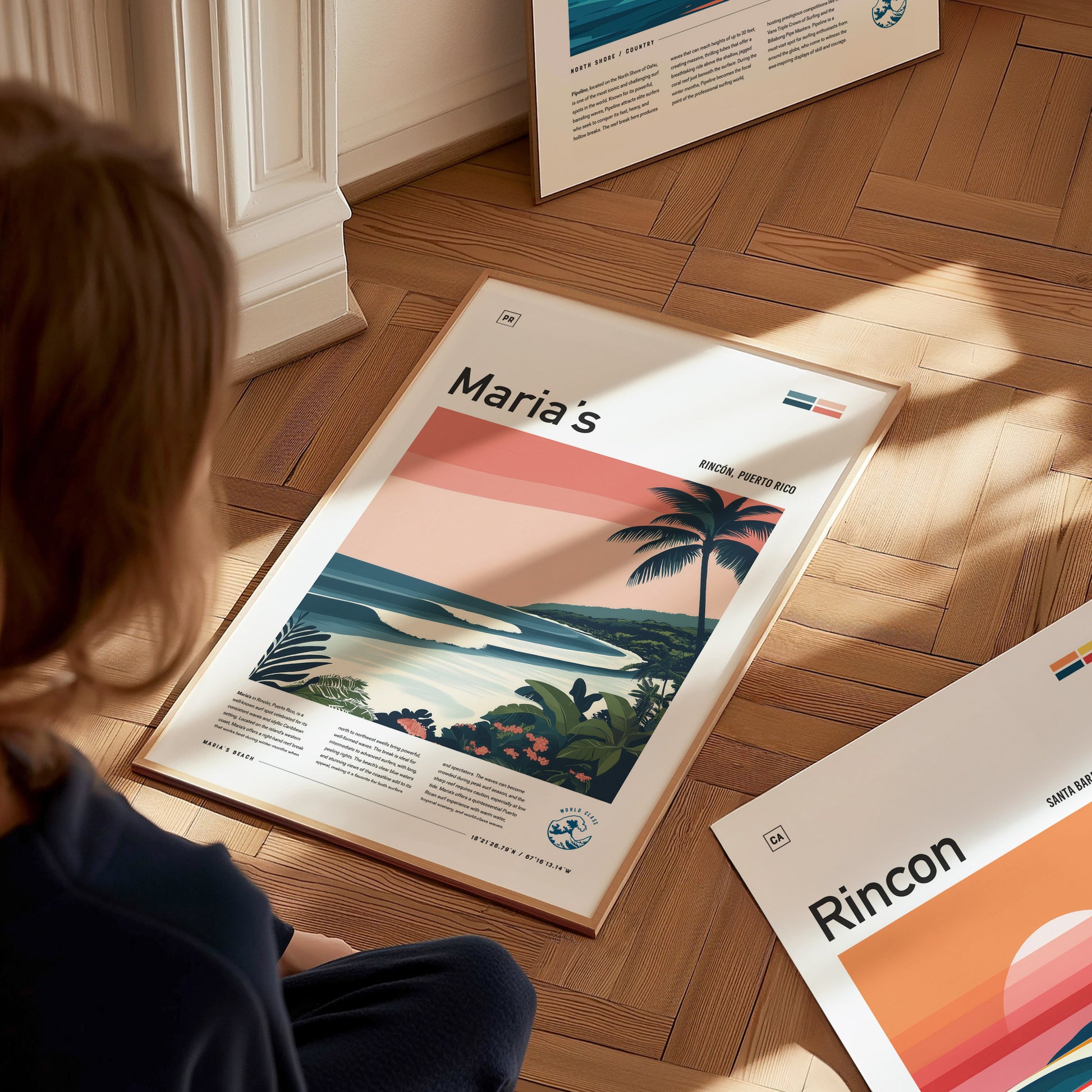 A person sits on a wooden floor, looking at a framed surf art print featuring a coastal scene with waves and palm trees labeled Marias. Other posters are visible nearby.