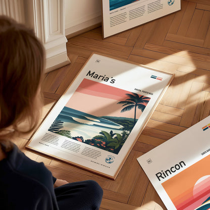 A person sits on a wooden floor, looking at a framed surf art print featuring a coastal scene with waves and palm trees labeled Marias. Other posters are visible nearby.