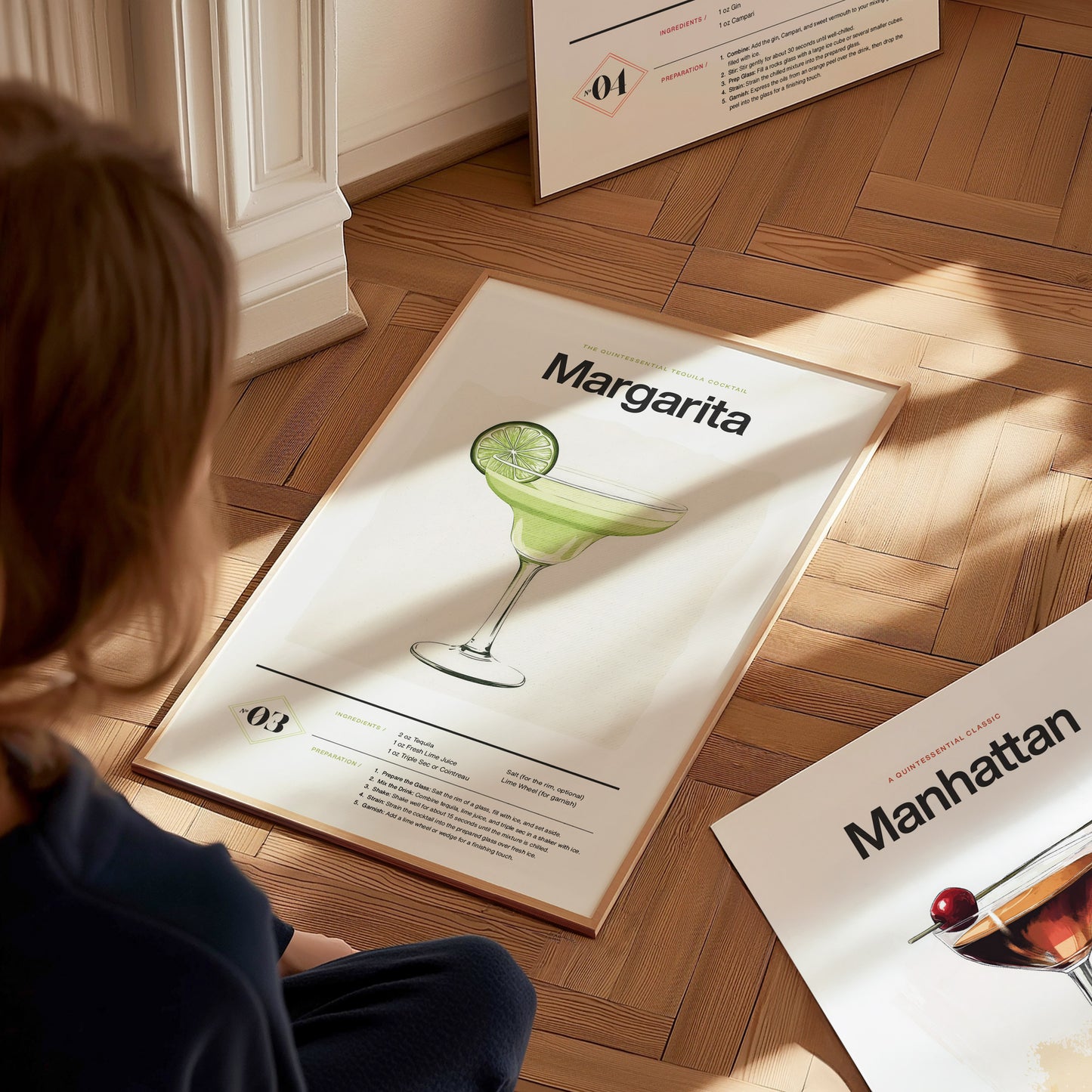 A person looks at a framed Margarita cocktail poster on a wooden floor, featuring a lime-garnished glass illustration. Another poster, partially visible, displays a Manhattan cocktail.
