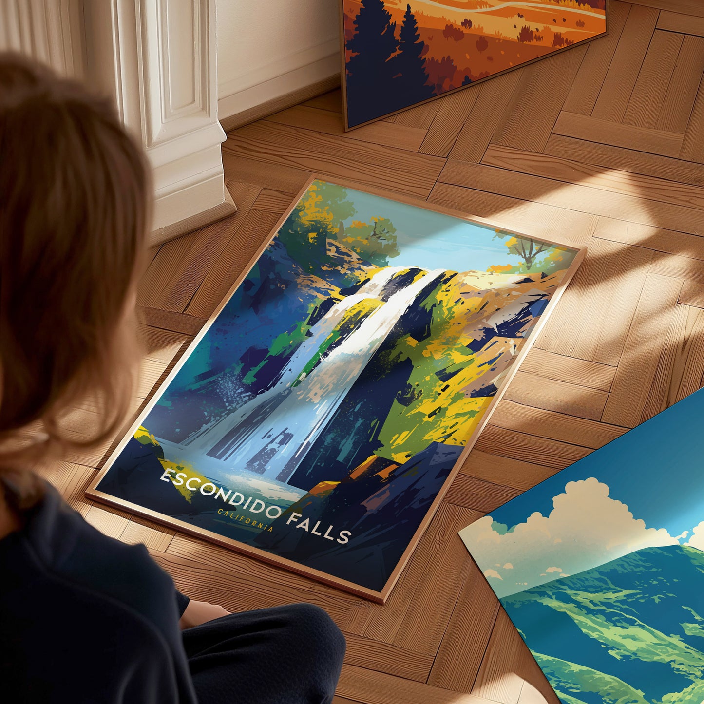 A person sits on a wooden floor, admiring a vibrant Escondido Falls poster featuring a waterfall and lush greenery. Sunlight casts soft shadows on the scene.