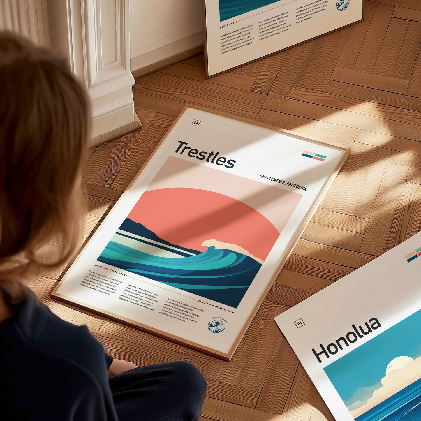 A person is sitting on a wooden floor, looking at a framed Trestles surf poster featuring stylized waves and a sunset. Other framed artworks are visible nearby.