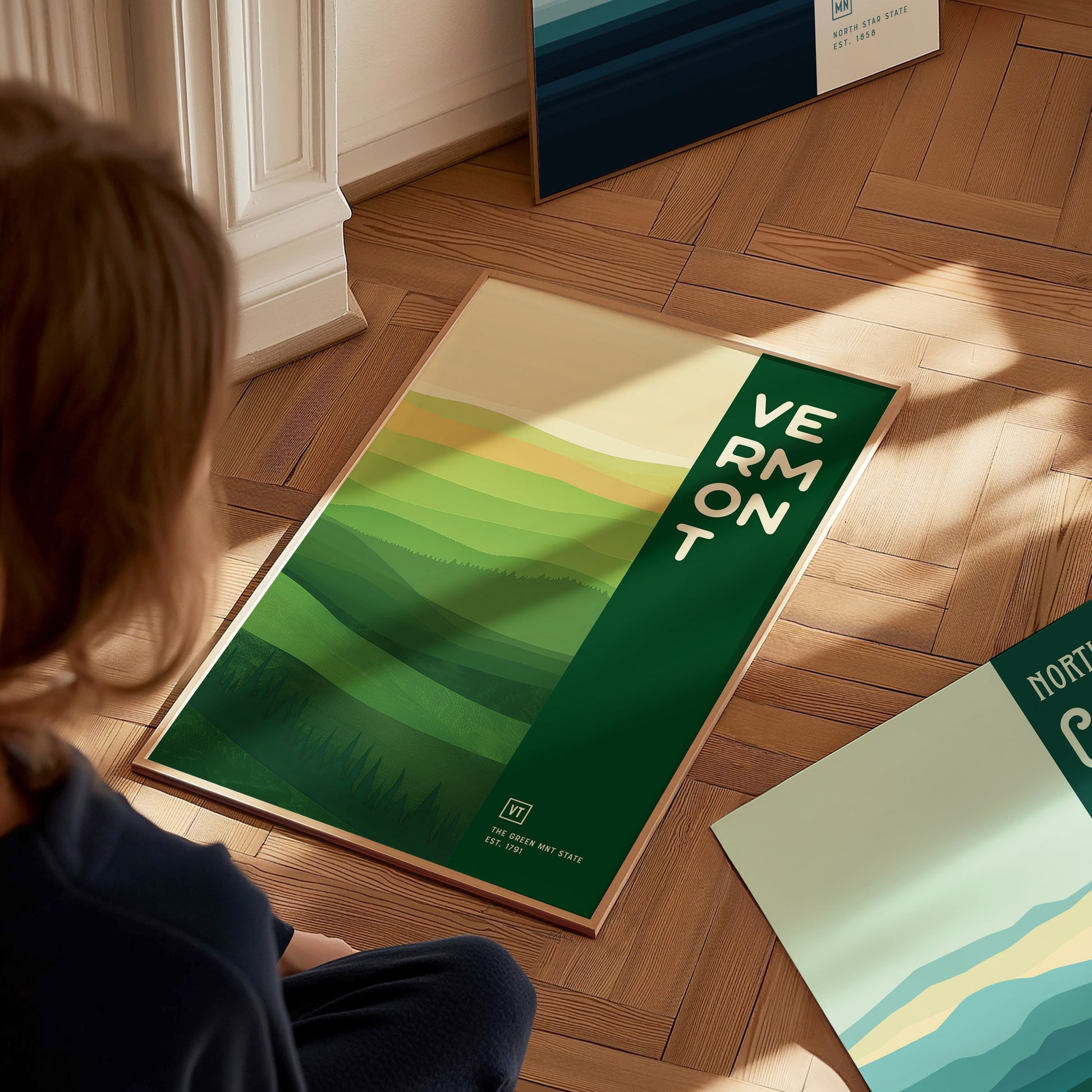 A person sits on the floor looking at a vibrant Vermont poster featuring abstract green hills and bold typography. Sunlight casts shadows on the wooden floor.