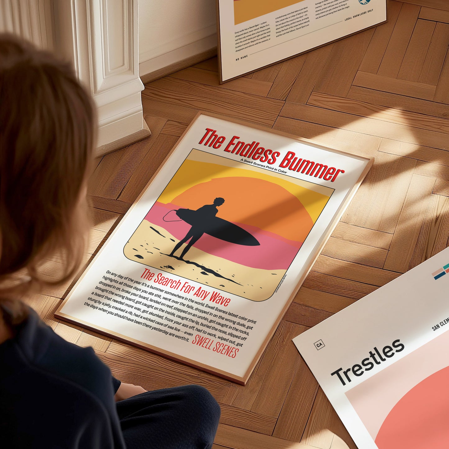 A person looks at a poster titled The Endless Bummer featuring a silhouetted surfer against a colorful sunset. Other posters are partially visible on the wooden floor.