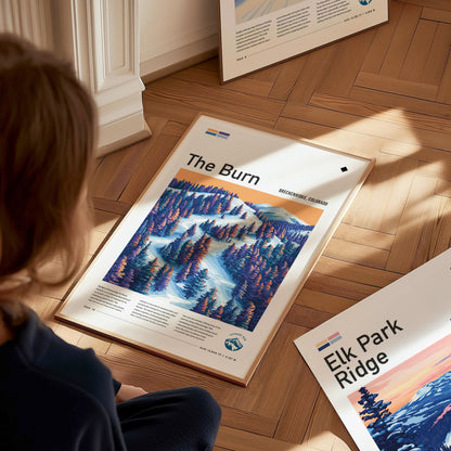 A person sits on a wooden floor, looking at a framed poster titled The Burn featuring a colorful ski run scene. Another poster, Elk Park Ridge, is partially visible.
