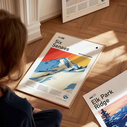 A person looks at a colorful poster titled Six Senses featuring a snowy mountain scene with vibrant sunset hues, lying on a wooden floor. Other posters are partially visible nearby.