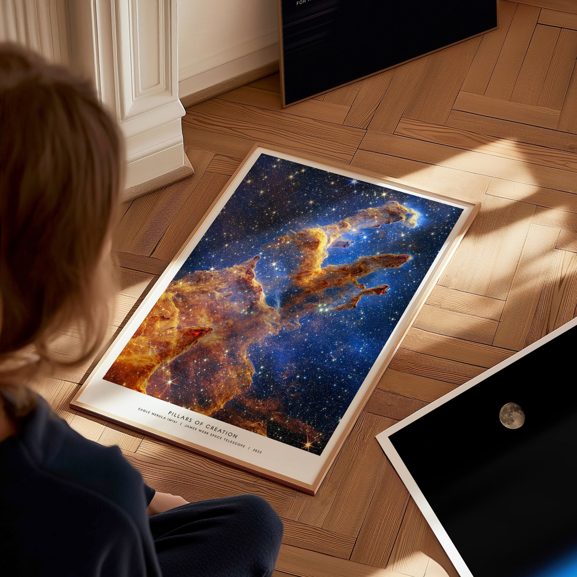 Large framed Pillars of Creation JWST artwork laying on a wooden floor beside other space prints, viewed from above.