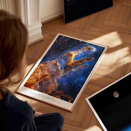 Large framed Pillars of Creation JWST artwork laying on a wooden floor beside other space prints, viewed from above.