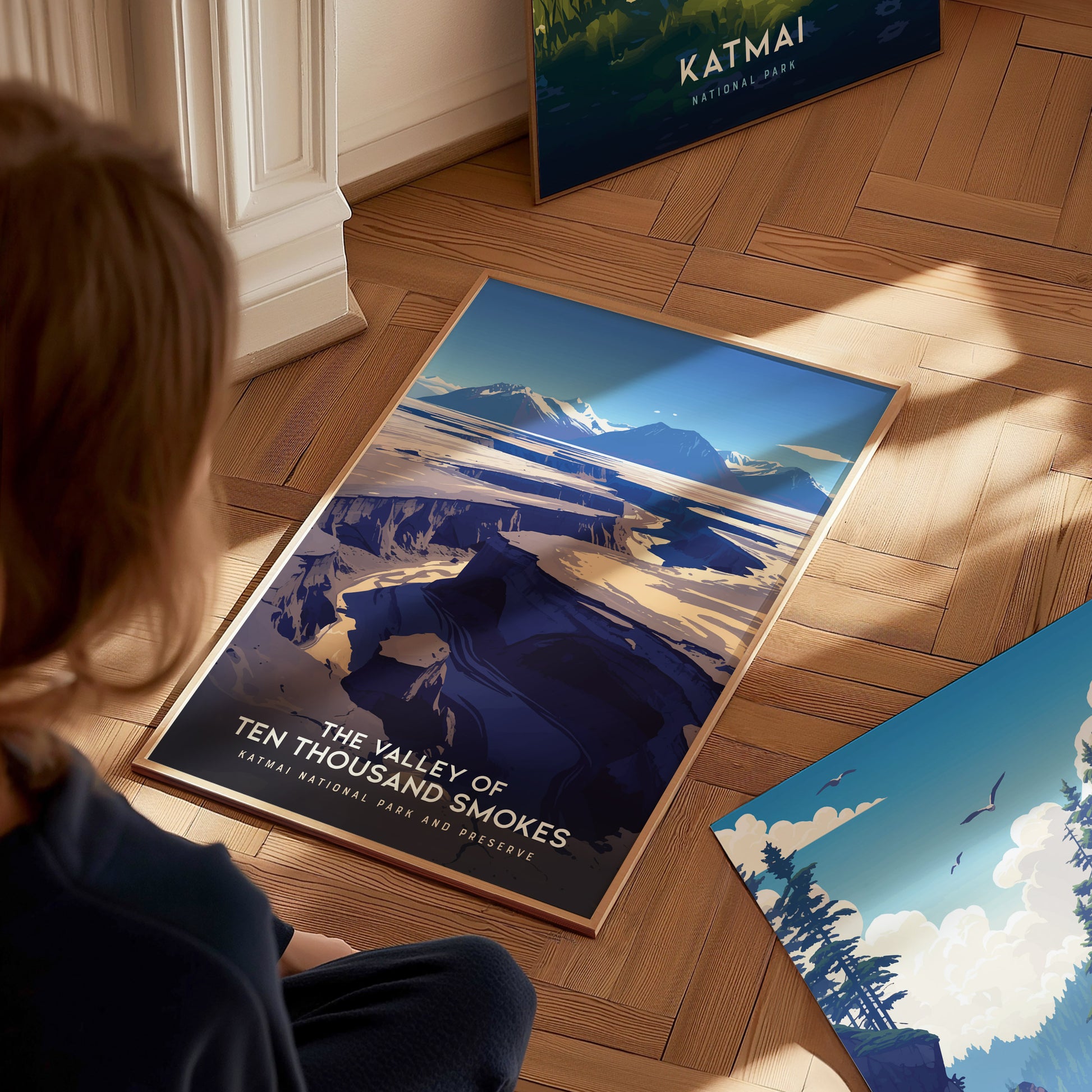 A person looks at a vibrant poster of the Valley of Ten Thousand Smokes, depicting a dramatic volcanic landscape with snowy peaks under a clear blue sky on a wooden.