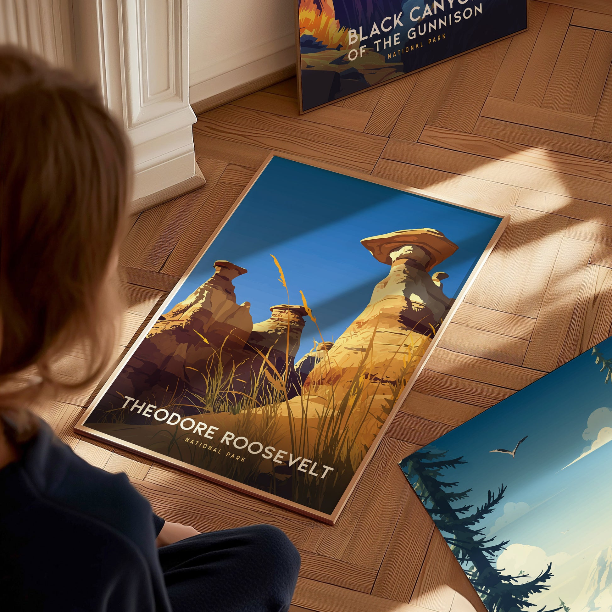 A person observes a Theodore Roosevelt National Park poster featuring striking hoodoos and grass against a blue sky, placed on a wooden floor. Another poster is partially visible nearby.