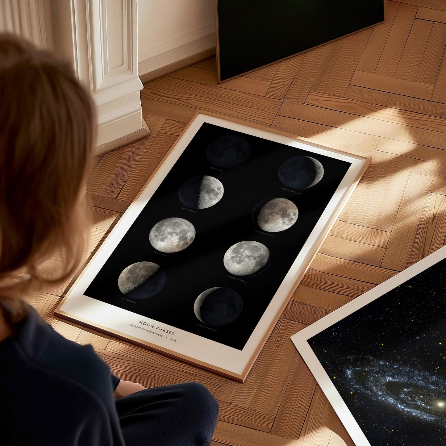 A child sits on a wooden floor, gazing at a framed poster displaying the eight phases of the Moon against a black background. The poster features detailed images of each lunar phase, highlighting the beauty of the lunar cycle.