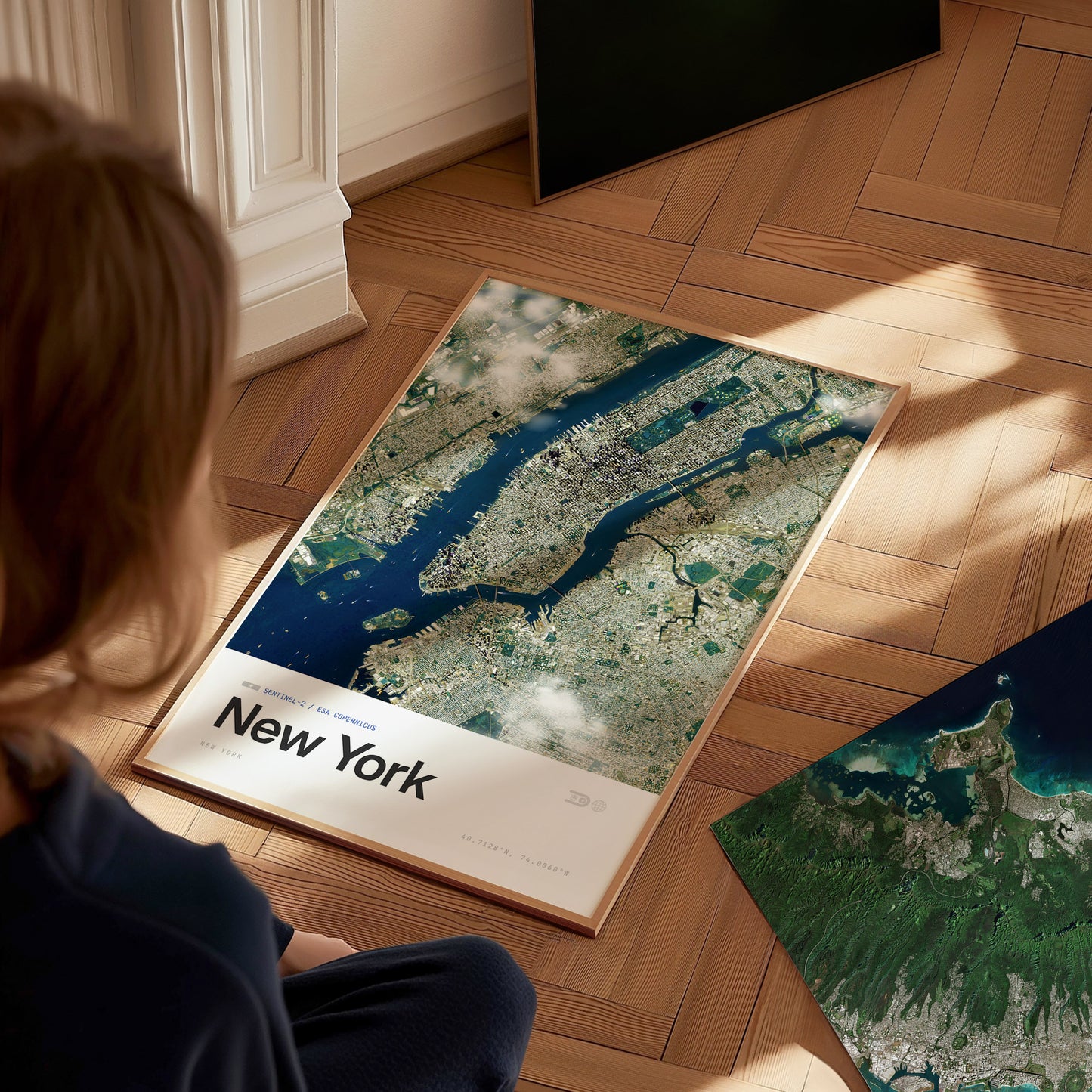 A child sits on a wooden floor, gazing at a framed aerial poster of New York City. The poster features a detailed satellite view of the city with its iconic waterways and urban landscape, showcasing vibrant colors and textures.