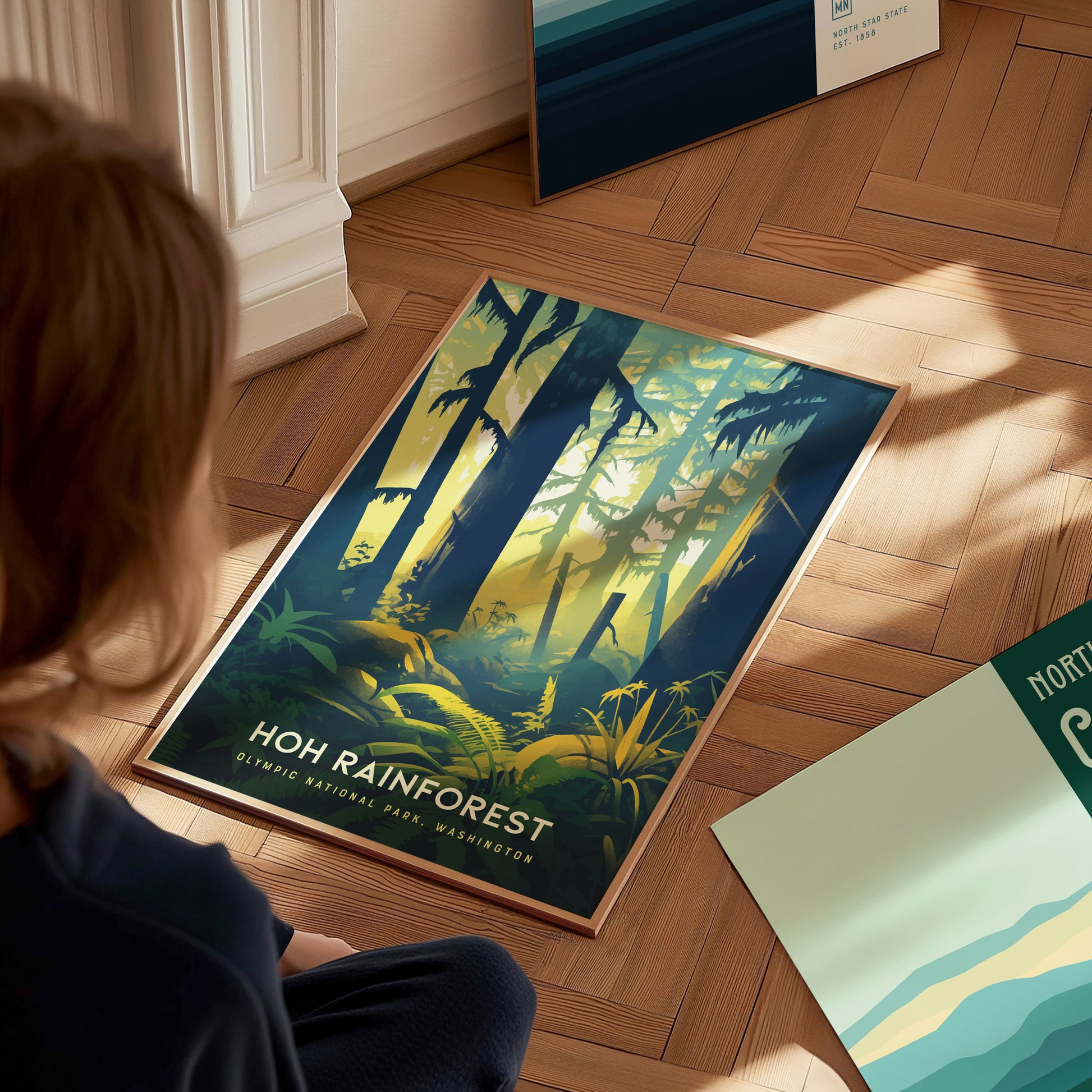 A person sits on a wooden floor, looking at a vibrant Hoh Rainforest poster featuring tall trees and lush greenery, with sunlight filtering through the forest canopy.