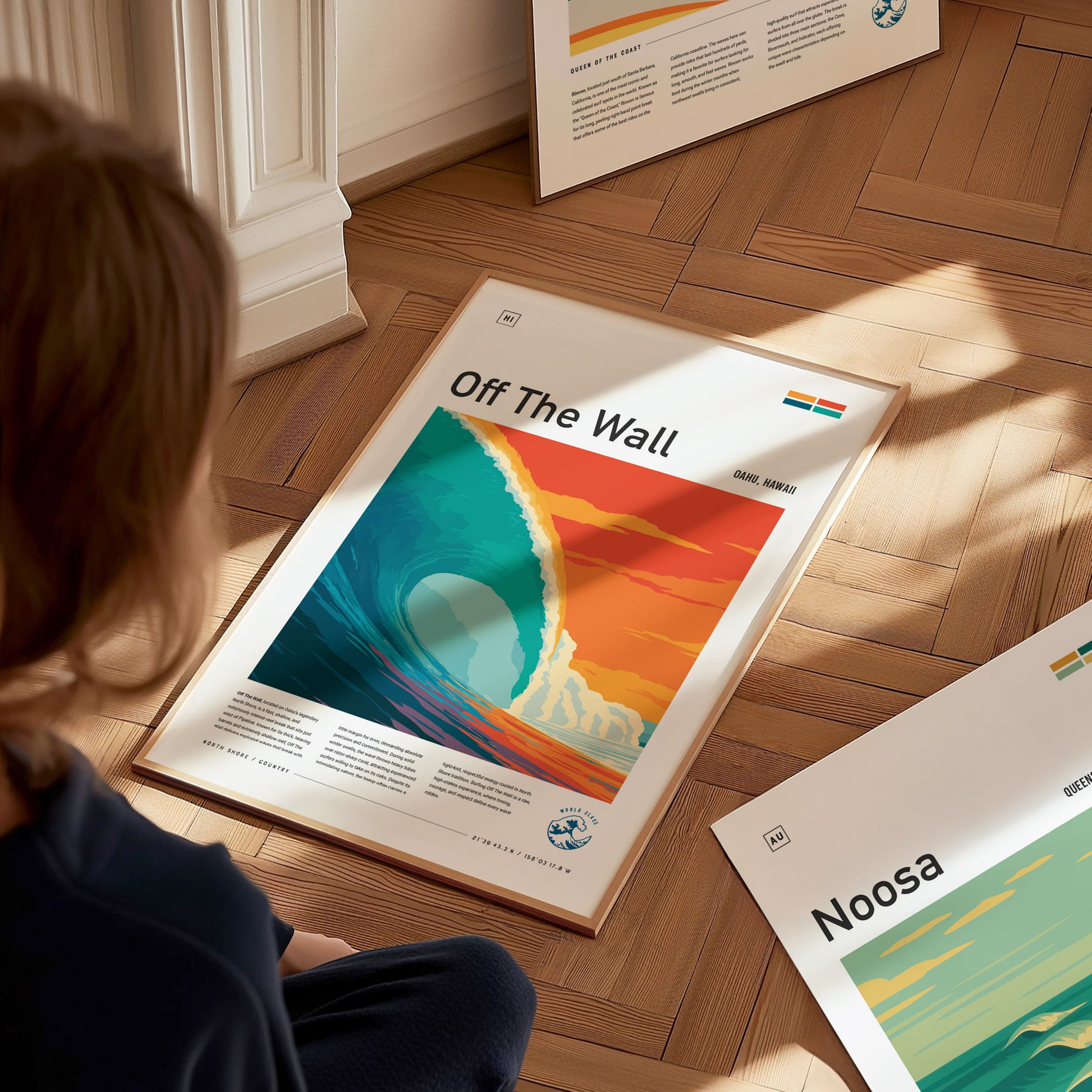 A child sits on a wooden floor, gazing at a vibrant art poster titled Off The Wall, featuring a large ocean wave in shades of teal and orange against a sunset backdrop.