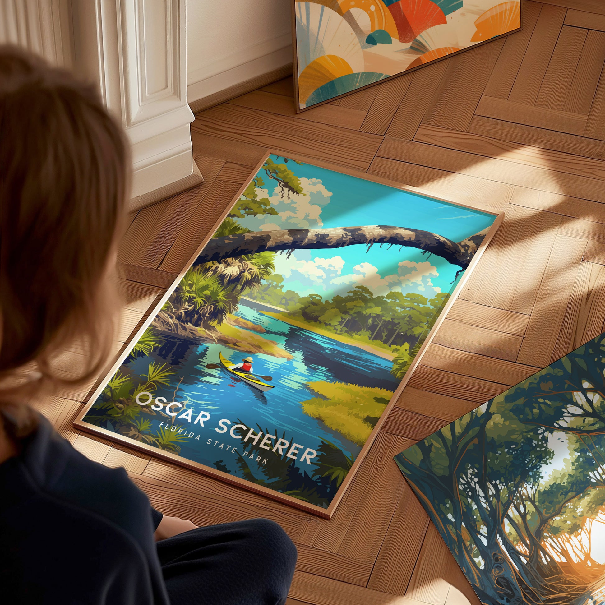 A person sits on a wooden floor, looking at a colorful poster of Oscar Scherer State Park featuring a canoeist on a serene river.