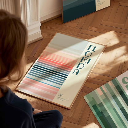 A person sits on a wooden floor, looking at a minimalist poster featuring the word Florida and colorful horizontal stripes. Another poster with green tones is partially visible.