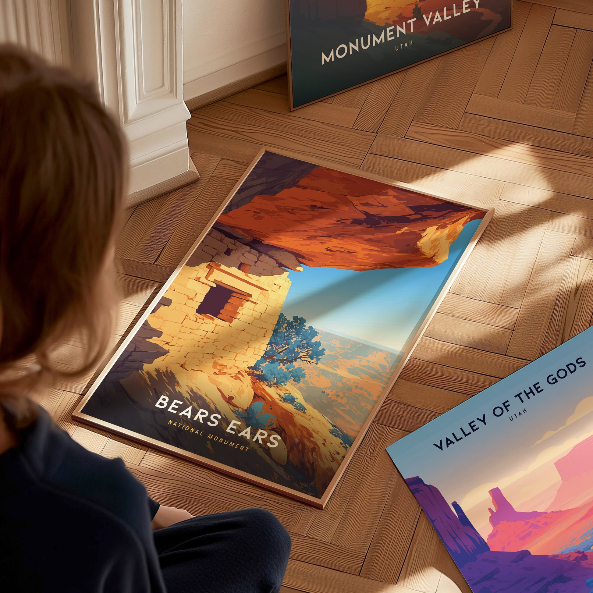 A person sits on a wooden floor looking at a Bears Ears National Monument poster, depicting vibrant desert scenery, with Monument Valley and Valley of the Gods posters nearby.